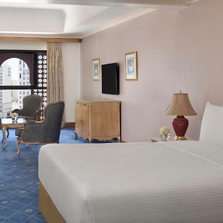 Hotel Intercontinental Dar Al Iman Madinah By Ihg Medina Esterno foto The photo shows a well-furnished hotel room. It features a large bed with a white duvet, two bedside lamps, and decorative paintings on the walls. There is a comfortable seating area with two chairs and a small table. A wooden dresser is positioned a