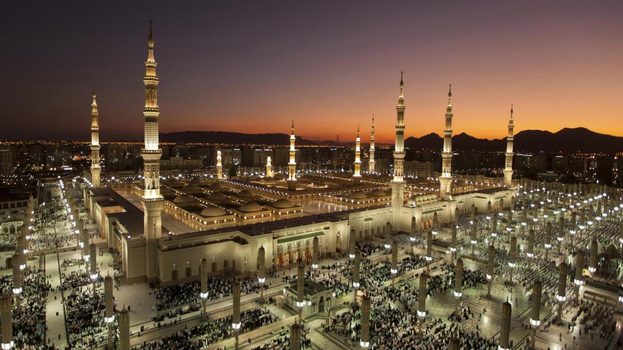 Hotel Intercontinental Dar Al Iman Madinah By Ihg Medina Esterno foto The photo depicts an aerial view of a large mosque complex during sunset. The mosque features multiple tall minarets, which are intricately designed and illuminated, giving the scene a stunning appearance as the sky transitions from daylight to dusk.
