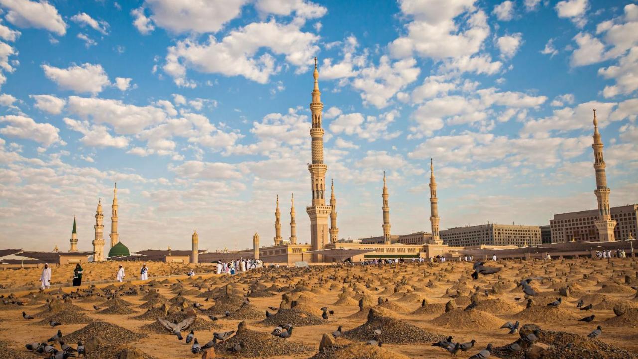 Hotel Intercontinental Dar Al Iman Madinah By Ihg Medina Esterno foto The photo shows a landscape that features a large mosque with a tall minaret surrounded by smaller minarets. In the foreground, there are numerous grave markers arranged in a desert-like setting. The sky is bright with scattered clouds, and there are