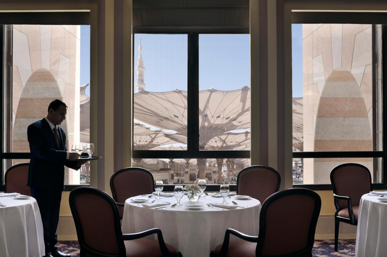 Hotel Intercontinental Dar Al Iman Madinah By Ihg Medina Esterno foto The photo shows a dining area with elegant decor. A waiter is standing to the side, holding a tray. The tables are neatly set with white tablecloths and fine dining arrangements. Large windows provide a view of an outdoor area featuring architectural