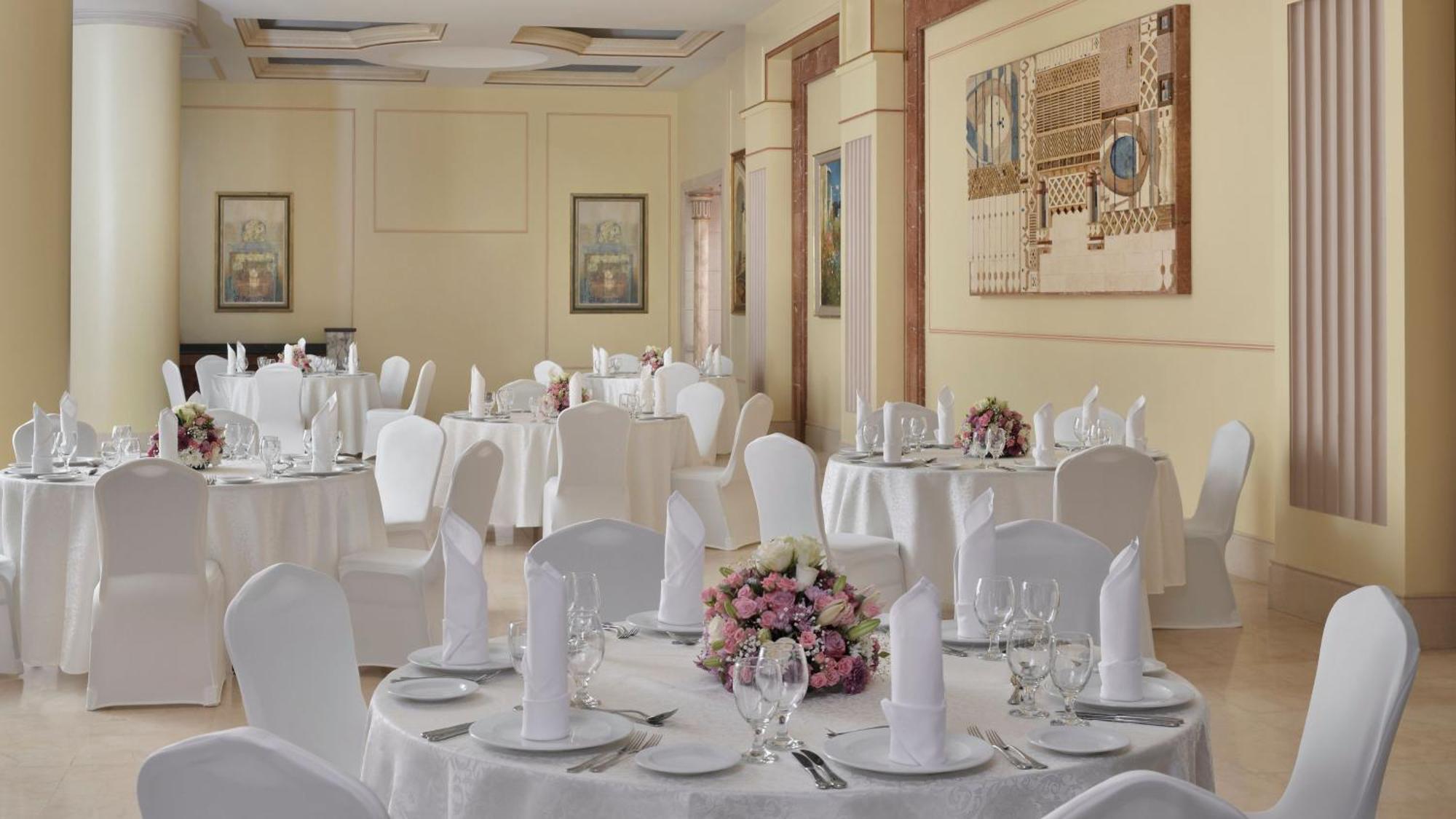Hotel Intercontinental Dar Al Iman Madinah By Ihg Medina Esterno foto The photo shows an elegantly arranged dining area, likely set up for a formal event or reception. There are several round tables covered with white tablecloths, each adorned with floral centerpieces featuring pink flowers. The tables are set with pla