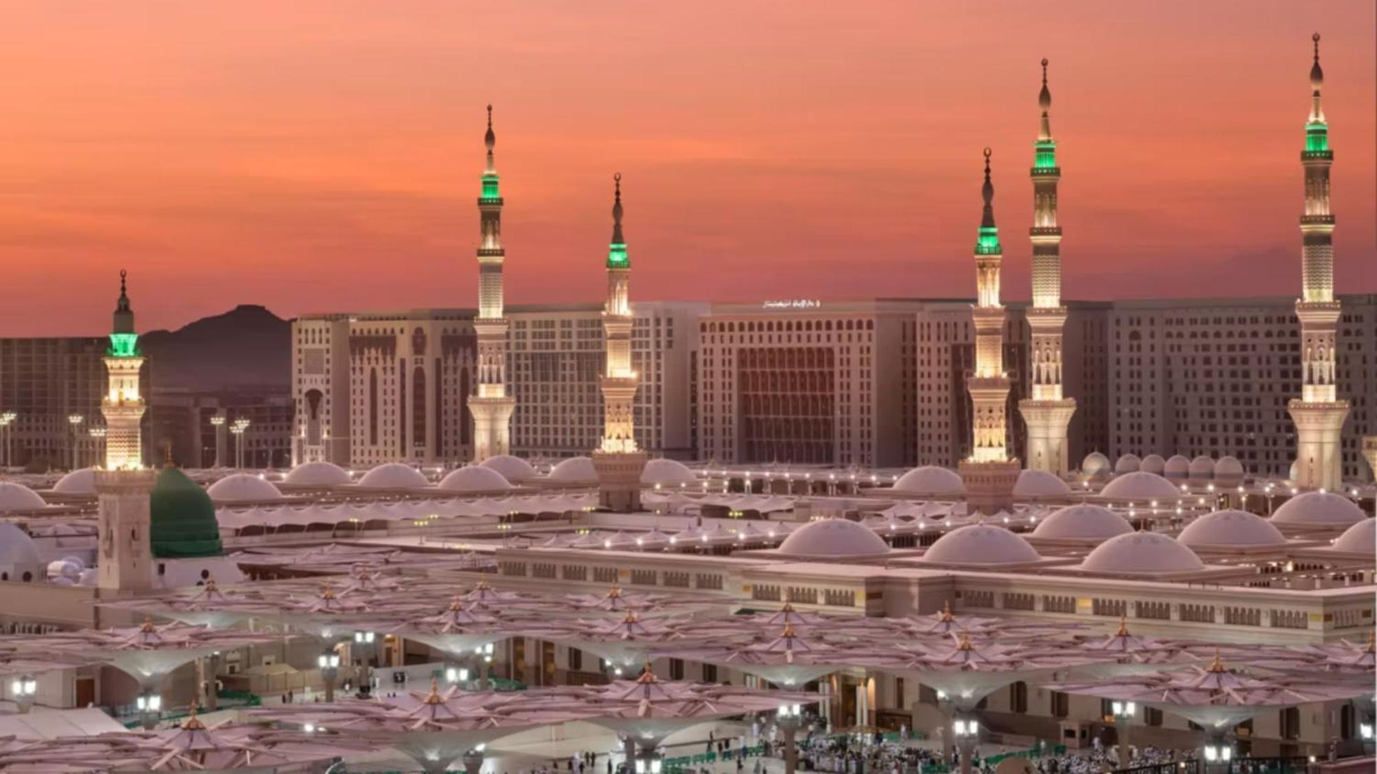 Hotel Intercontinental Dar Al Iman Madinah By Ihg Medina Esterno foto The photo depicts a scenic view of a mosque complex, likely during sunset, with several minarets illuminated and showcasing green lights. The architecture features domed roofs, and the setting includes a backdrop of buildings, all bathed in a warm, o