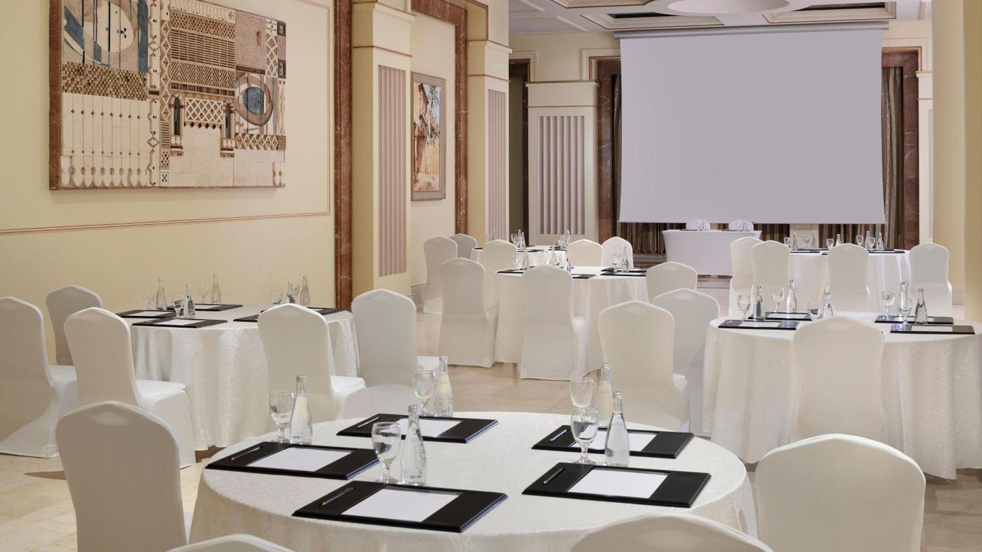 Hotel Intercontinental Dar Al Iman Madinah By Ihg Medina Esterno foto The photo depicts an elegantly arranged event space. There are several round tables covered with white tablecloths and surrounded by white chairs. Each table is set with a black folder or menu and a glass of water. The walls are adorned with artworks
