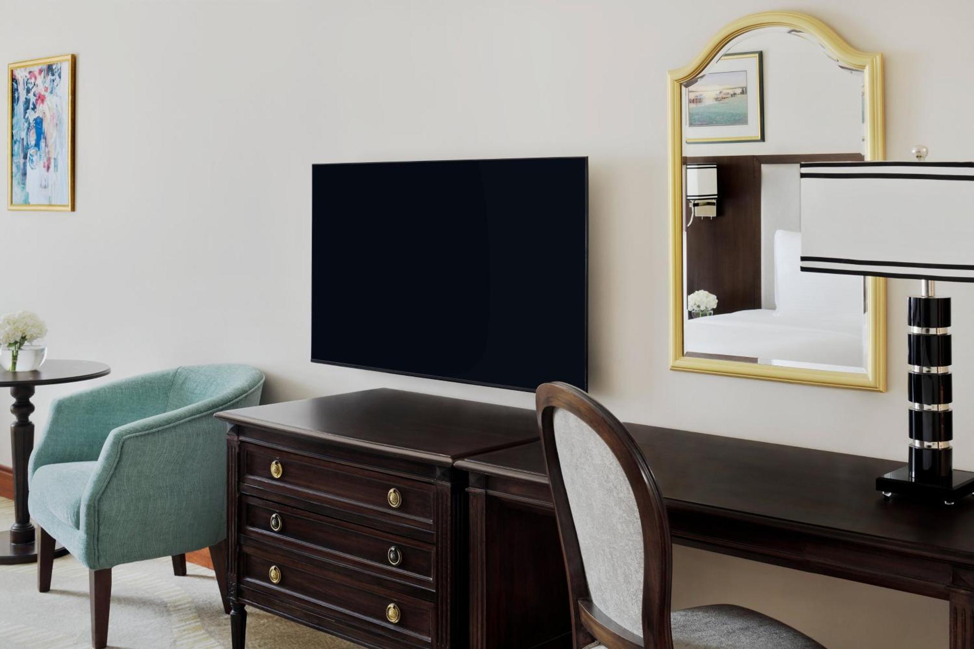 Hotel Intercontinental Dar Al Iman Madinah By Ihg Medina Esterno foto The photo shows a hotel room interior featuring a mounted flat-screen television on the wall. Below the TV, there is a dark wooden dresser with drawers. To the right, there's a wooden desk and a chair with a light-colored seat. A small table with a l