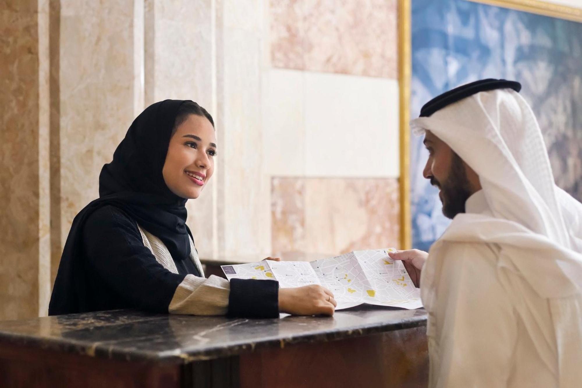Hotel Intercontinental Dar Al Iman Madinah By Ihg Medina Esterno foto The photo depicts a woman in a black hijab and a light-colored outfit smiling as she interacts with a man wearing traditional Emirati attire, including a white kandura and ghutrah (headscarf). They are at a reception desk in what appears to be a hote