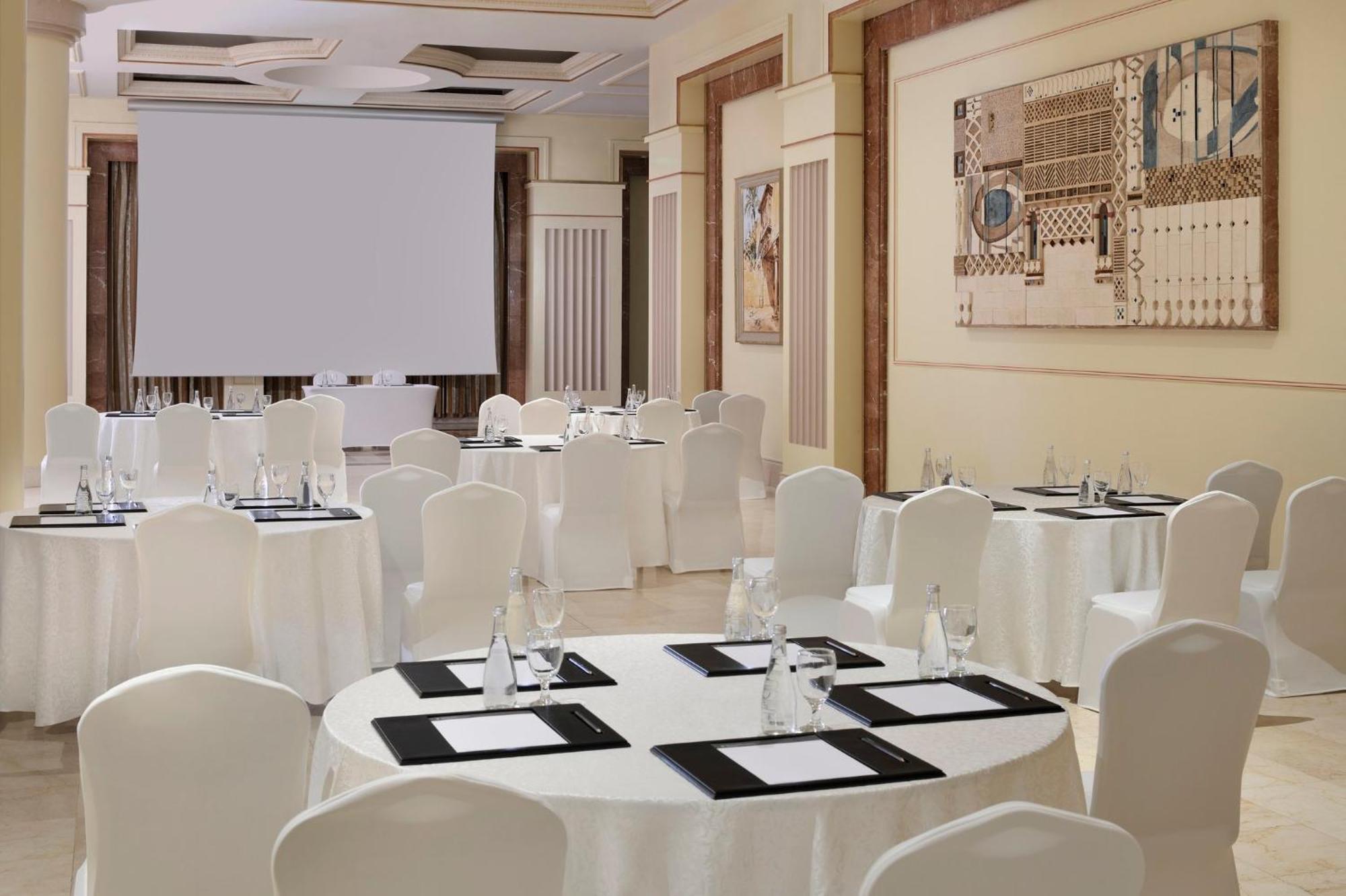 Hotel Intercontinental Dar Al Iman Madinah By Ihg Medina Esterno foto The photo shows a well-decorated event space or meeting room. It features several round tables covered with white tablecloths and surrounded by white chairs. Each table has black placemats and glass water bottles, suggesting a formal setup for a gath