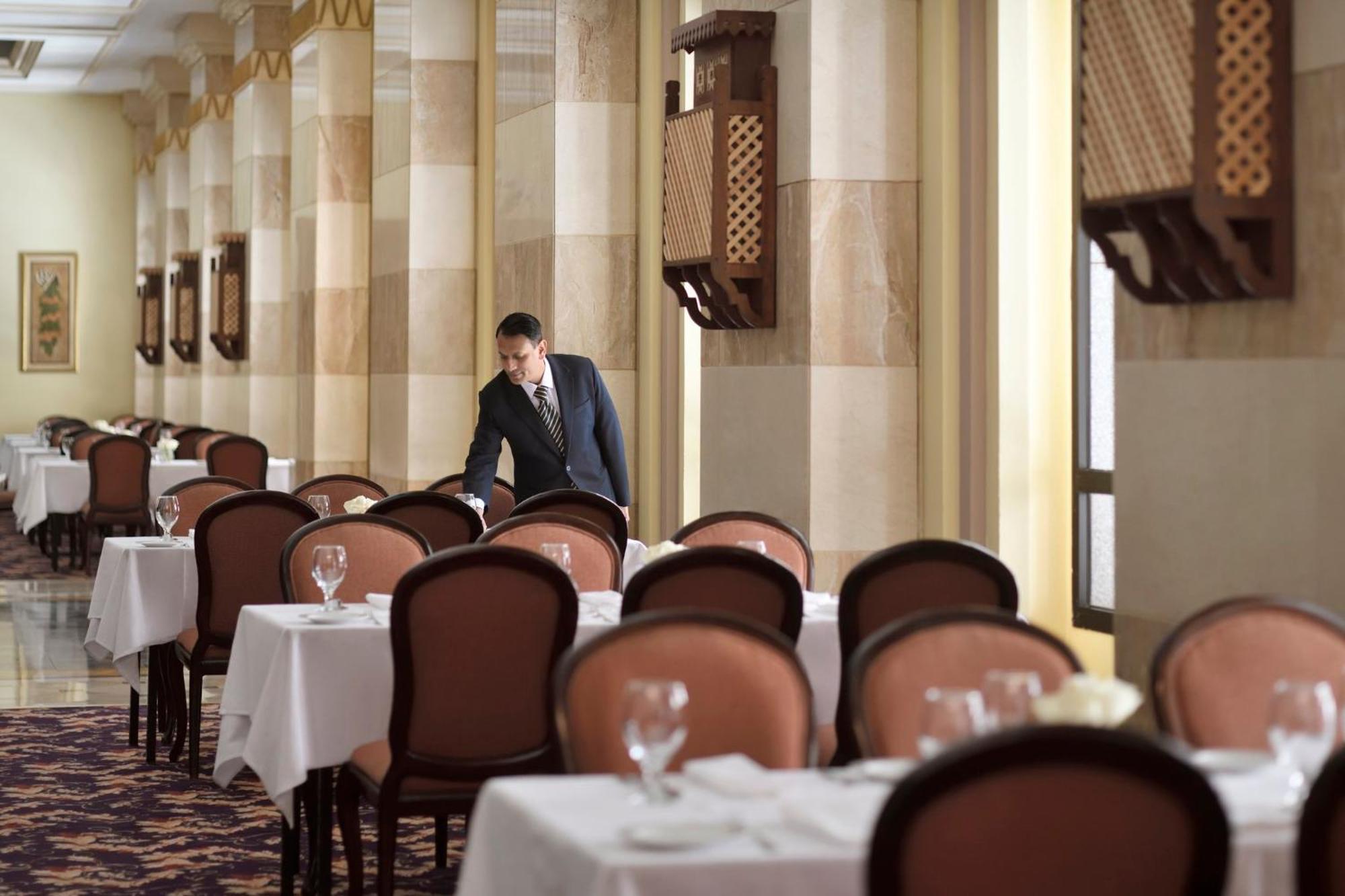 Hotel Intercontinental Dar Al Iman Madinah By Ihg Medina Esterno foto The photo depicts a dining area in an elegant restaurant. There are several tables set with white tablecloths, each accompanied by chairs. A waiter, dressed in a formal suit, is seen arranging or tending to one of the tables. The restaurant features 