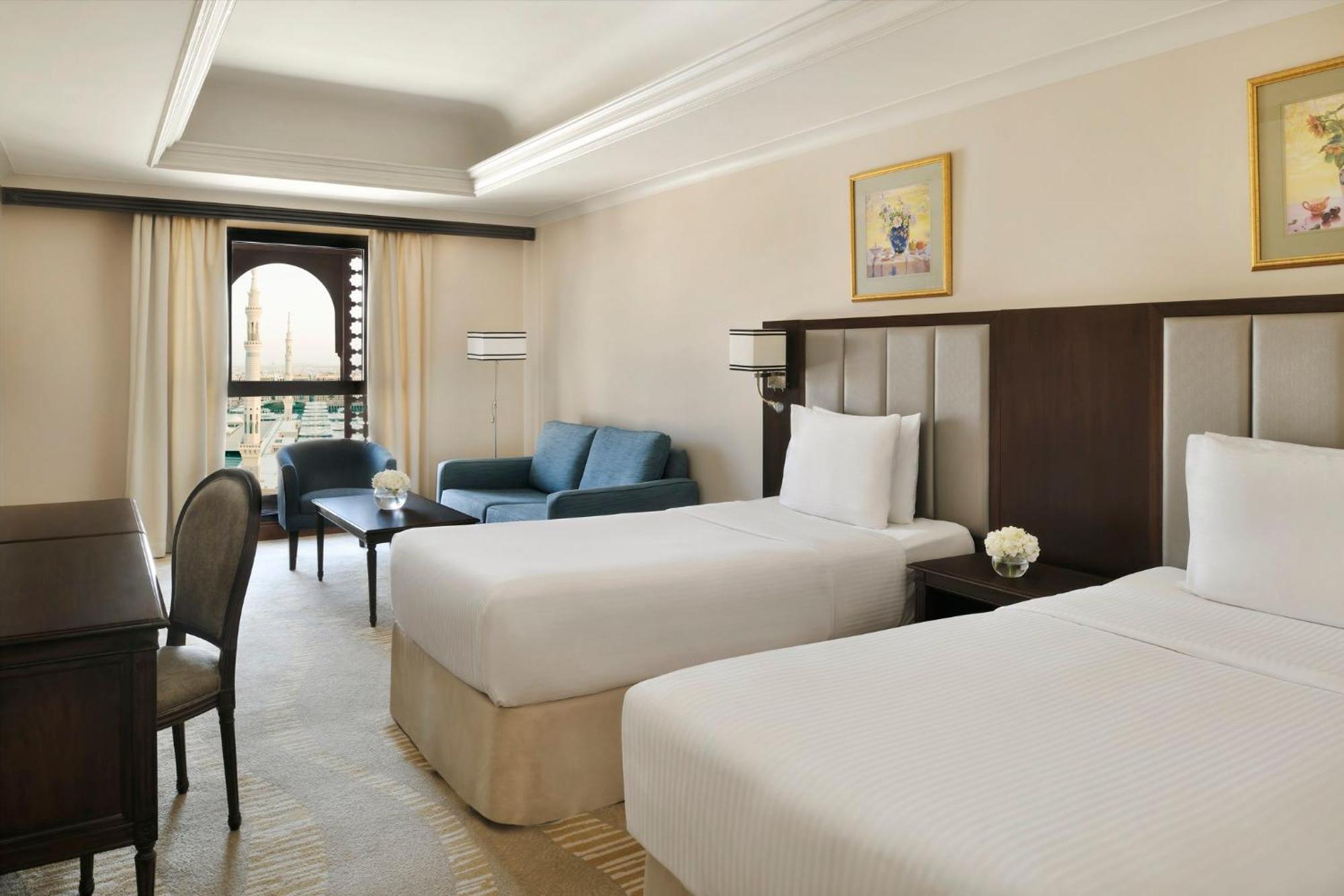 Hotel Intercontinental Dar Al Iman Madinah By Ihg Medina Esterno foto The photo shows a well-appointed hotel room featuring two twin beds with white bedding. There is a small desk with a chair, a comfortable blue sofa, and a coffee table in the sitting area. The room has a warm, neutral color scheme, accented by two pa
