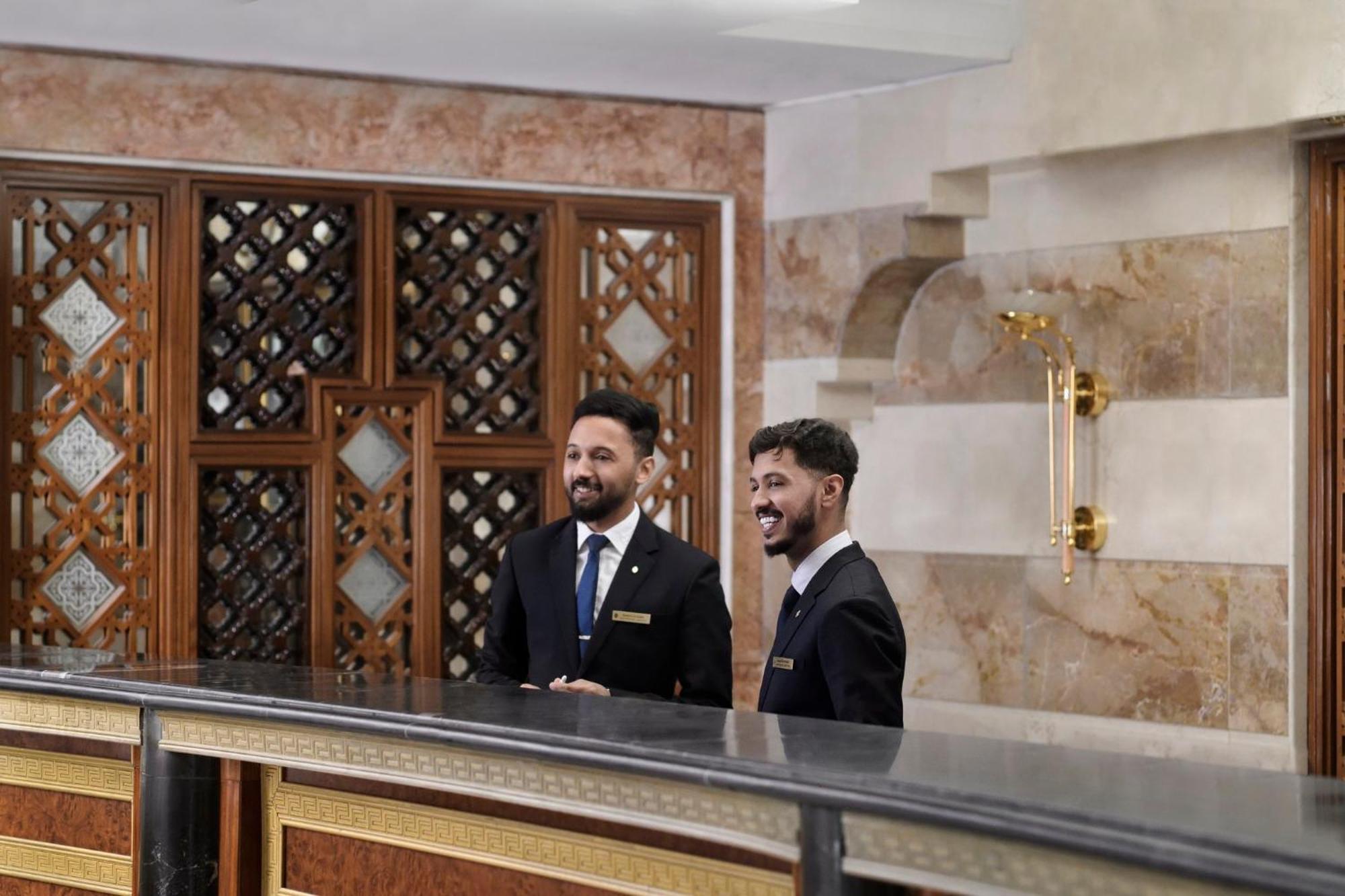 Hotel Intercontinental Dar Al Iman Madinah By Ihg Medina Esterno foto The photo shows two men standing behind a reception desk in an elegant setting, likely a hotel or similar establishment. They are both dressed in formal attire, wearing suits and ties, and appear to be smiling or engaging in conversation. The backgro