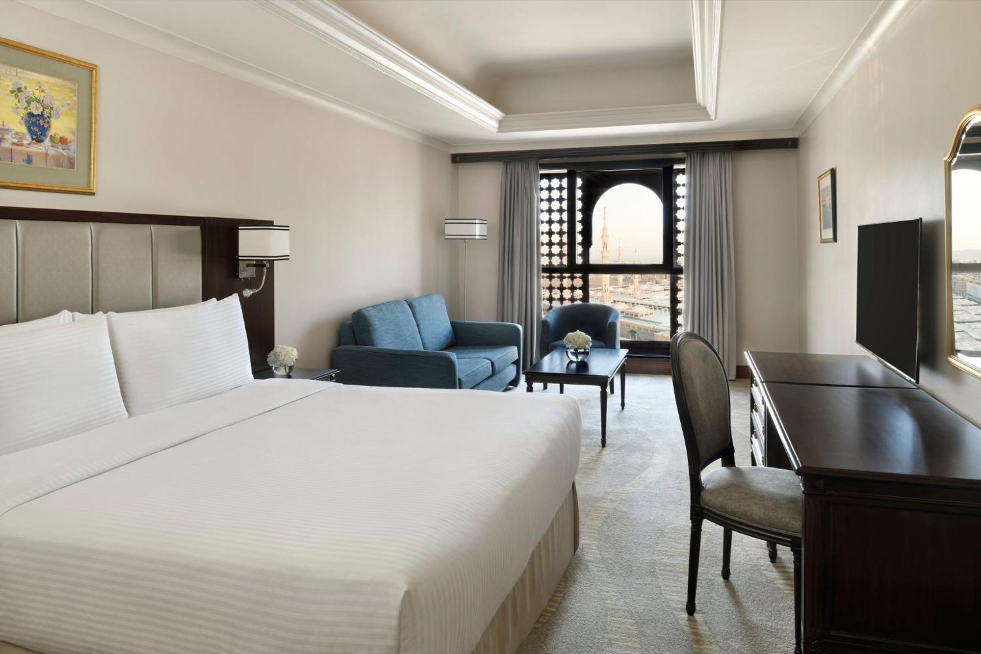 Hotel Intercontinental Dar Al Iman Madinah By Ihg Medina Esterno foto The photo shows a hotel room designed with a modern and comfortable aesthetic. It features a large bed with white linens, a seating area with a blue sofa, and a small table. There is a wooden desk with a chair and a television opposite the bed. Natur
