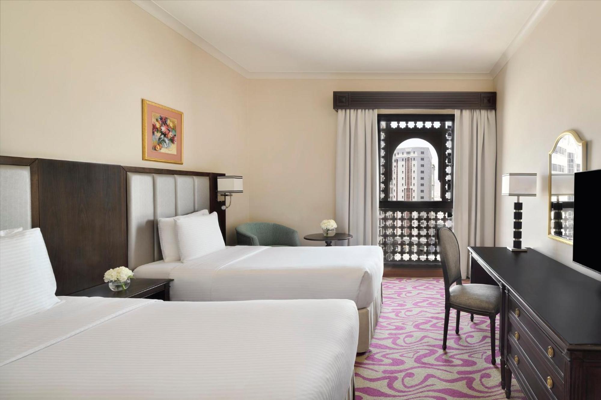 Hotel Intercontinental Dar Al Iman Madinah By Ihg Medina Esterno foto The photo shows a hotel room featuring two twin beds with white bedding. There is a decorative piece of art on the wall above one of the beds. A small round table with a flower arrangement is placed next to the beds. A desk and chair are situated aga
