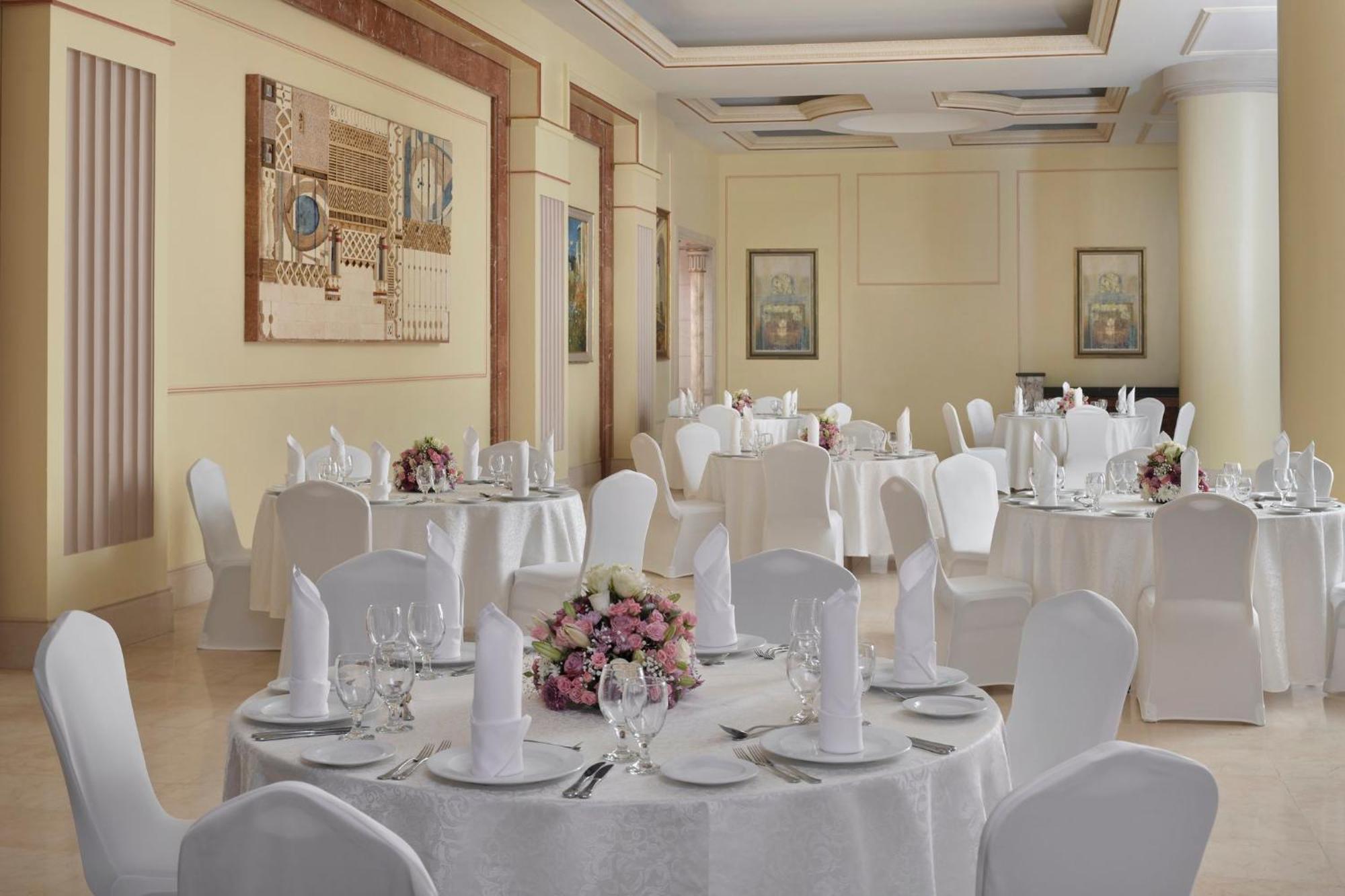 Hotel Intercontinental Dar Al Iman Madinah By Ihg Medina Esterno foto The photo shows an elegantly arranged dining or function room. There are round tables covered with white tablecloths, each set with fine dining ware including glassware, utensils, and neatly folded white napkins. Some tables have floral centerpieces 