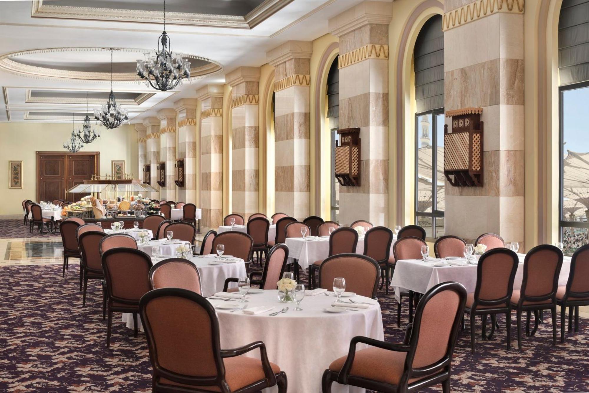 Hotel Intercontinental Dar Al Iman Madinah By Ihg Medina Esterno foto The photo depicts an elegant dining room setting. It features several round tables covered with white tablecloths, each set with dishes and glassware. The room has a sophisticated ambiance, characterized by high ceilings adorned with chandeliers and 