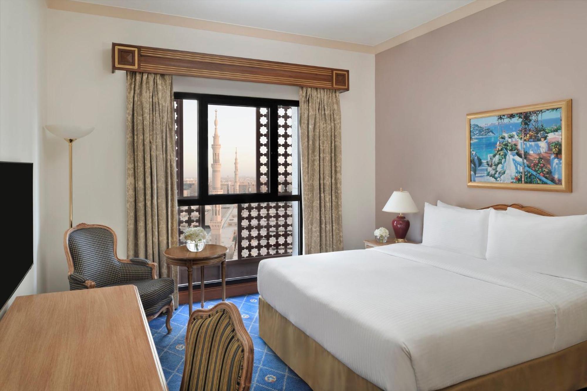 Hotel Intercontinental Dar Al Iman Madinah By Ihg Medina Esterno foto The photo shows a hotel room with a modern design. There is a large bed with white linens placed against the wall. Next to the bed, there is a small bedside table with a lamp. A comfortable armchair is situated nearby, accompanied by a wooden desk. T