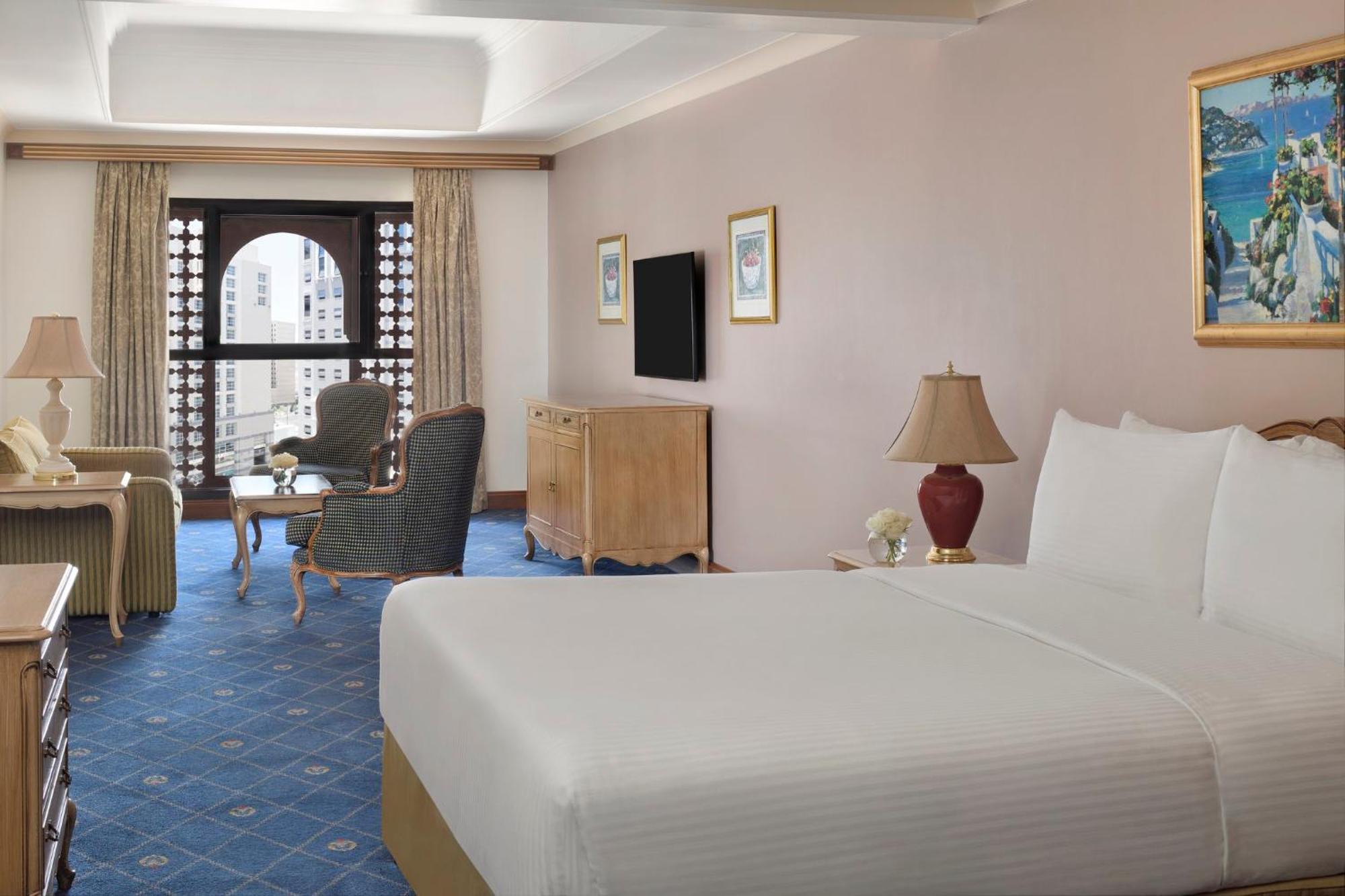 Hotel Intercontinental Dar Al Iman Madinah By Ihg Medina Esterno foto The photo shows a well-furnished hotel room. It features a large bed with a white duvet, two bedside lamps, and decorative paintings on the walls. There is a comfortable seating area with two chairs and a small table. A wooden dresser is positioned a