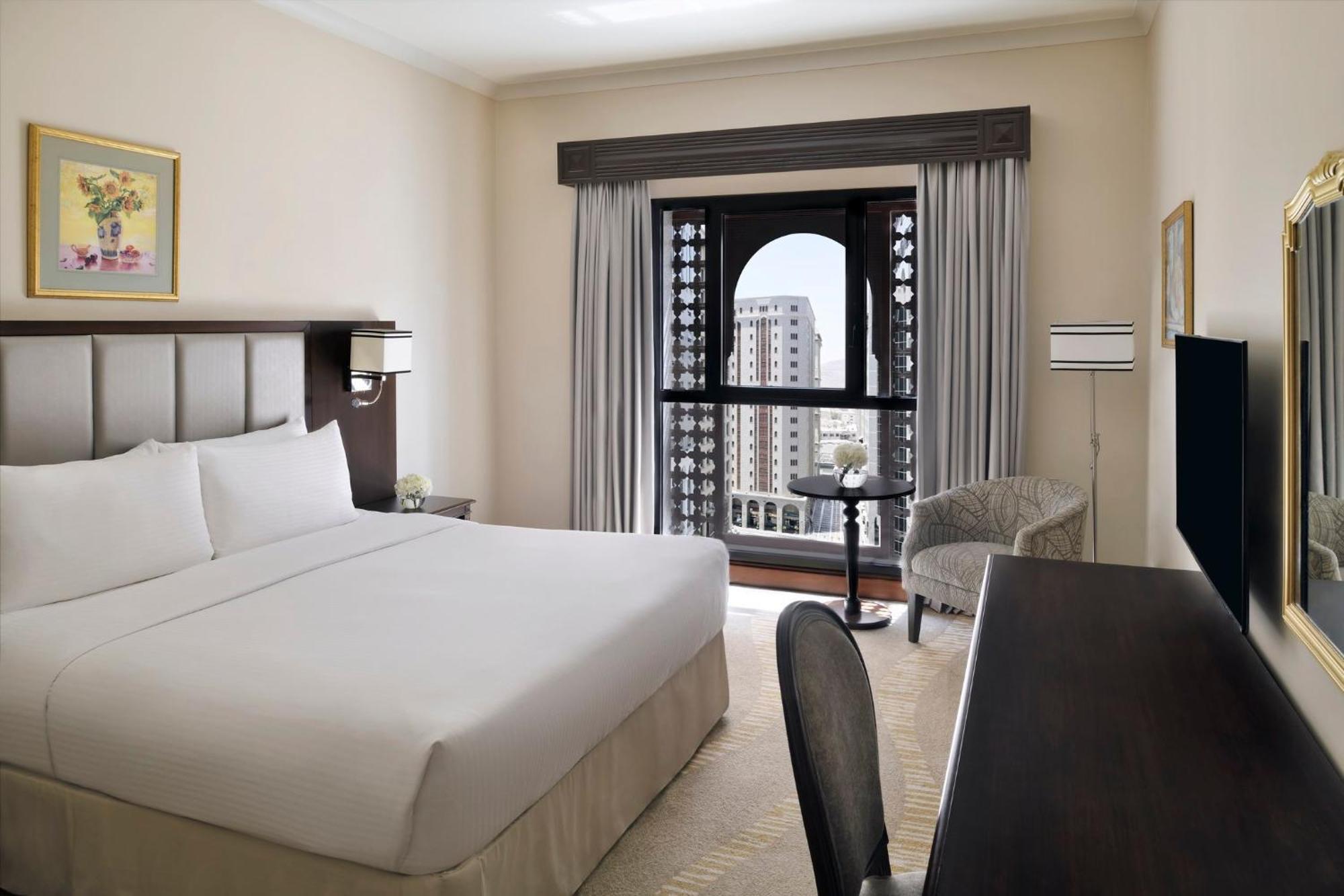 Hotel Intercontinental Dar Al Iman Madinah By Ihg Medina Esterno foto The photo shows a hotel room with a modern and minimalistic design. It features a large bed with white linens, a side table, and a comfortable chair near a window. The window has a view of a cityscape, and elegant curtains frame the window. There is 