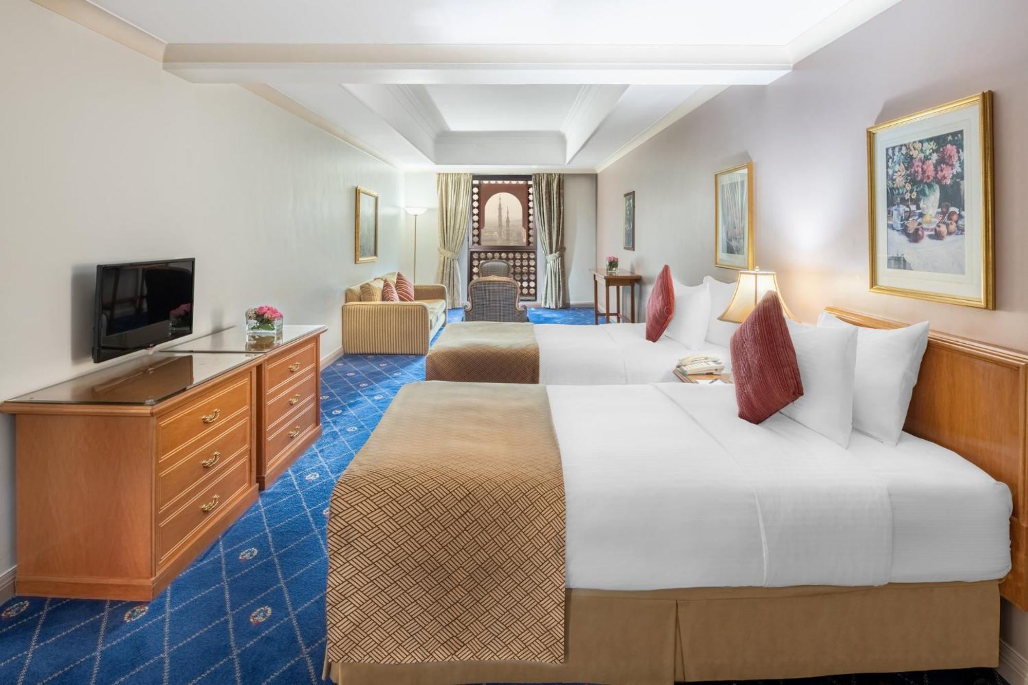 Hotel Intercontinental Dar Al Iman Madinah By Ihg Medina Esterno foto The photo shows a hotel room featuring two beds with white linens and decorative pillows. There is a wooden dresser with a television on it. The flooring has a blue carpet with a pattern, and the walls are painted in a light color. The room is well-l