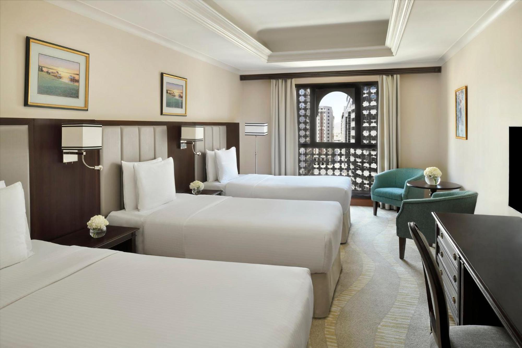Hotel Intercontinental Dar Al Iman Madinah By Ihg Medina Esterno foto The photo shows a hotel room with three single beds, dressed in white linens. There are two paintings on the walls, depicting serene landscapes. A large window allows natural light to enter, and it is adorned with elegant curtains. A smaller seating 