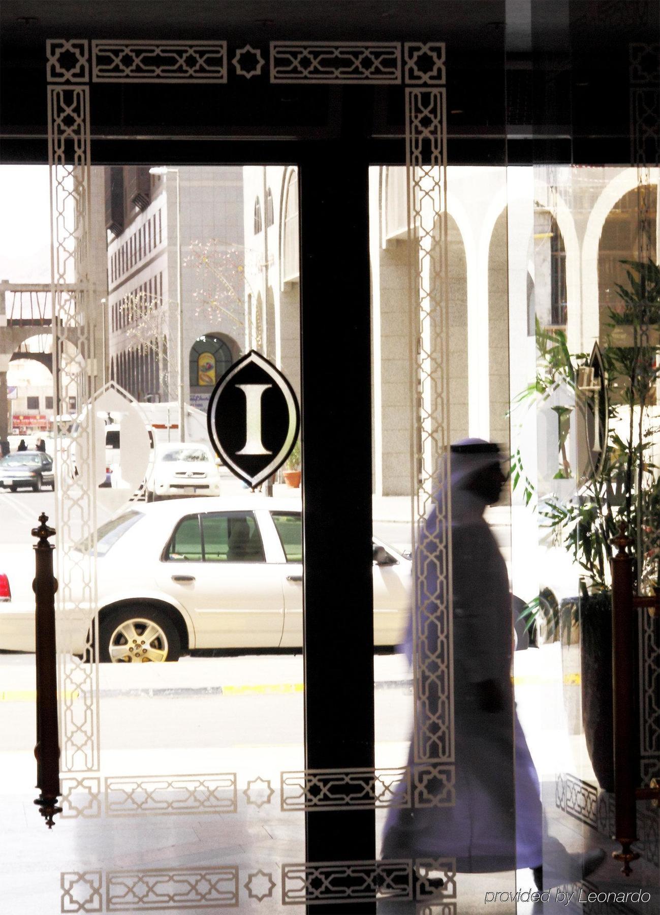 Hotel Intercontinental Dar Al Iman Madinah By Ihg Medina Interno foto The photo shows a view through a glass door or window, featuring an elegant design with a prominent "I" symbol displayed. Through the glass, there are some parked vehicles visible, including a white car. In the foreground, a shadowy figure of a perso