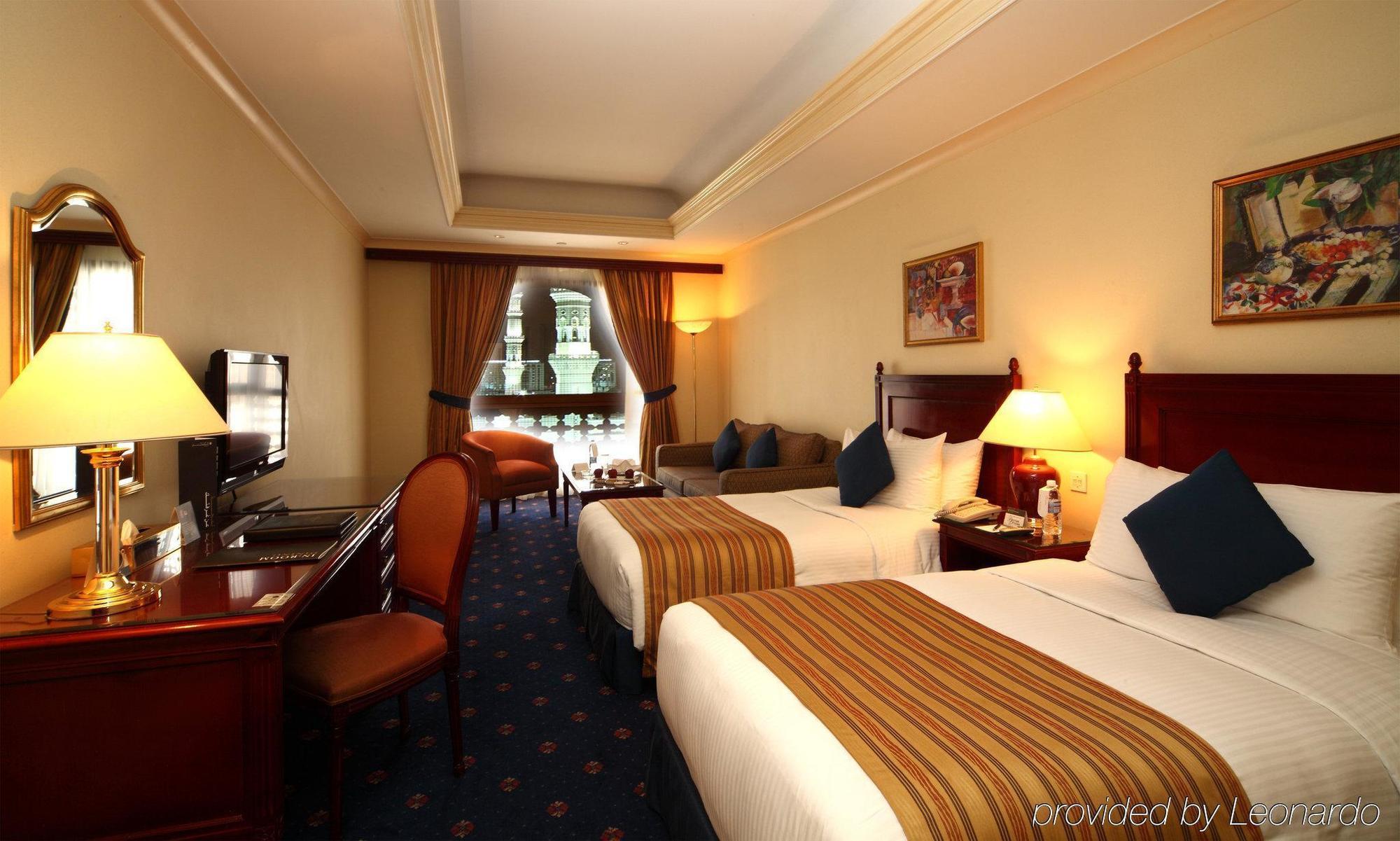 Hotel Intercontinental Dar Al Iman Madinah By Ihg Medina Camera foto The photo shows a hotel room featuring two beds with white linens and brown striped throws. There is a wooden desk and chair, alongside a television. The walls are painted in a warm tone, and there are decorative paintings. Large windows with curtain
