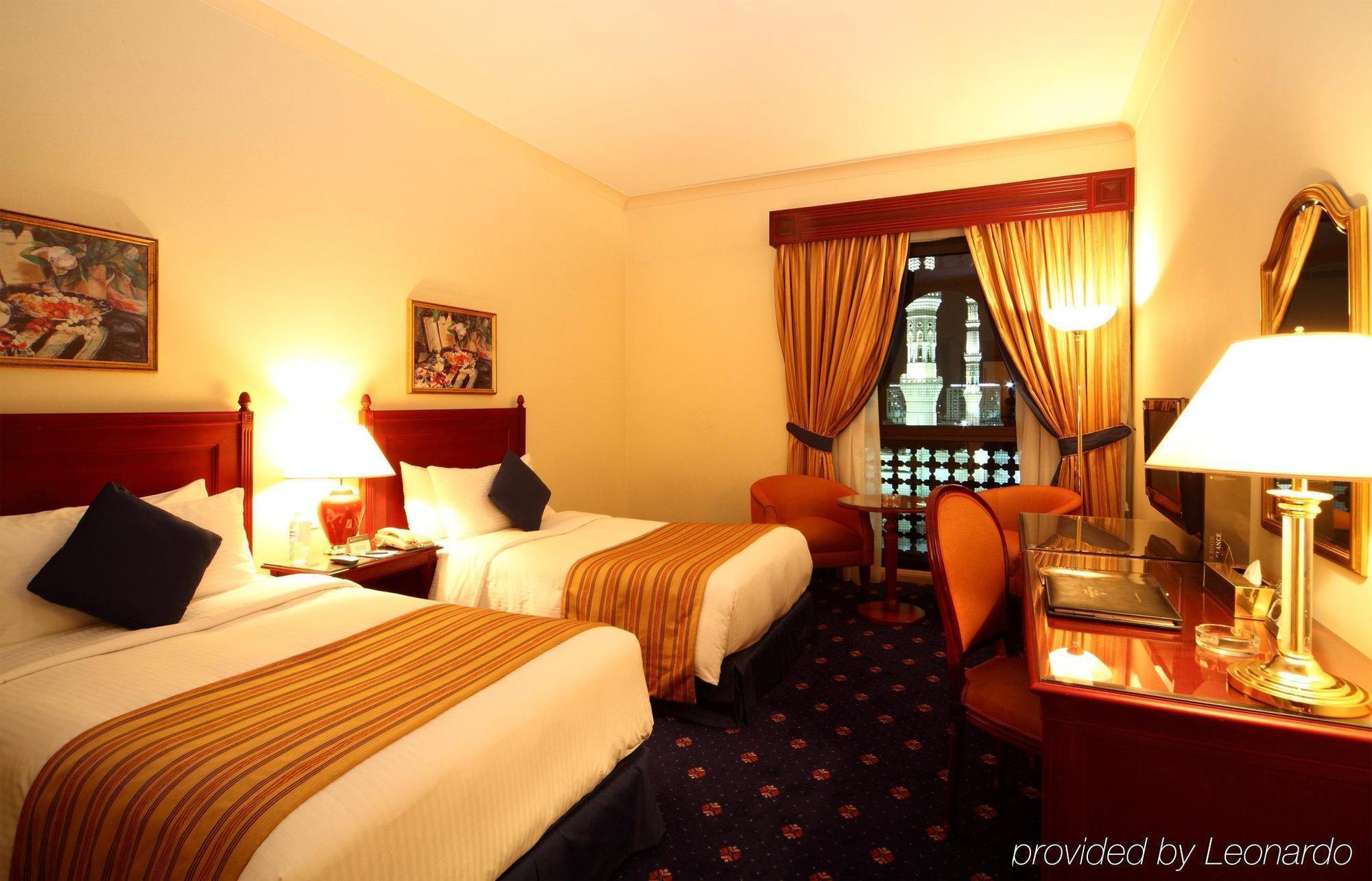 Hotel Intercontinental Dar Al Iman Madinah By Ihg Medina Camera foto The photo shows a hotel room featuring two twin beds with white linens and brown striped bedspreads. There are decorative pillows on the beds, and the room has warm-colored walls. To the side, there's a small sitting area with a round table and a cha