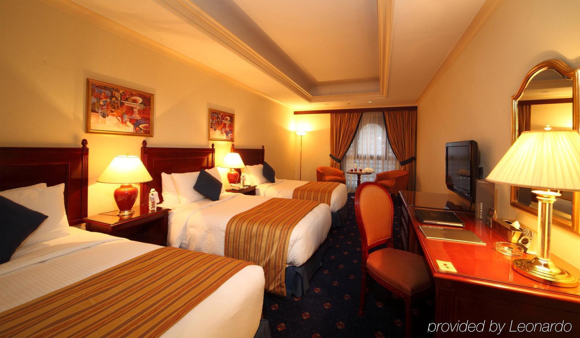 Hotel Intercontinental Dar Al Iman Madinah By Ihg Medina Camera foto The photo shows a hotel room with a warm and inviting atmosphere. There are two beds with white and gold bedding, complemented by decorative pillows. Each bed has a bedside lamp. In the background, there's a small seating area with a couple of chairs