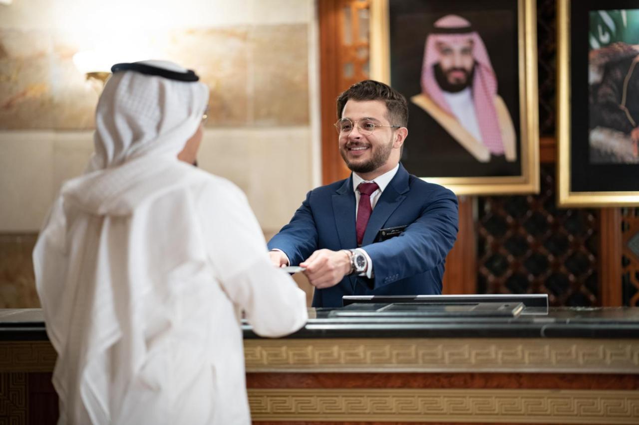 Hotel Intercontinental Dar Al Iman Madinah By Ihg Medina Esterno foto The photo shows a hotel reception area. A male receptionist, dressed in a navy suit with a tie, is smiling and handing an item to a guest who is wearing traditional white attire, possibly a kandura or thobe. The background features elegantly decorate