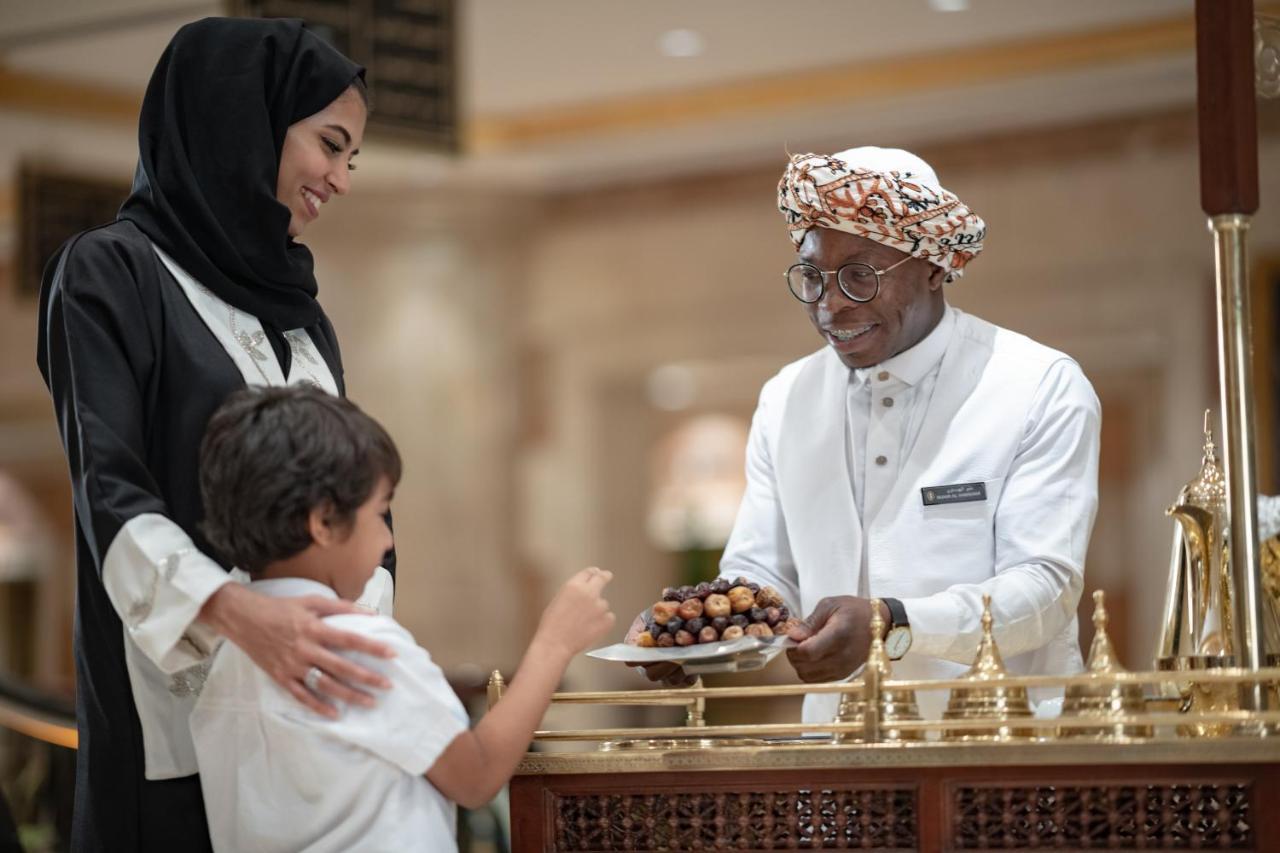 Hotel Intercontinental Dar Al Iman Madinah By Ihg Medina Esterno foto The photo depicts a warm interaction between three people. A woman, dressed in a traditional black attire and a headscarf, stands beside a young boy who is reaching out towards a plate of dates held by a smiling man in a white traditional outfit with